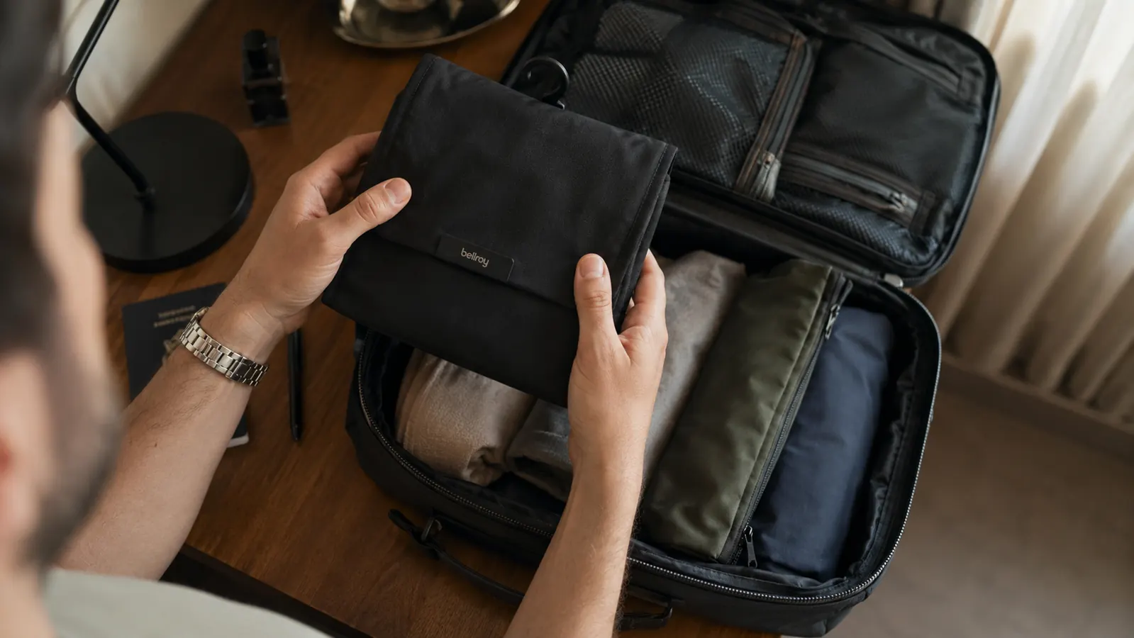 Traveler packing toiletry bag into a carry-on suitcase at a hotel room desk